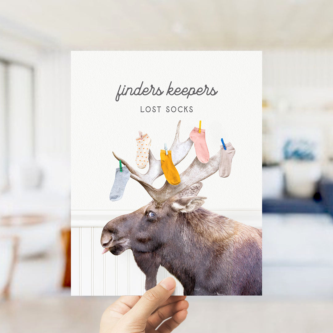 A hand holding a card with a moose wearing socks and antlers, with the text "finders keepers" and "lost socks" written on it.