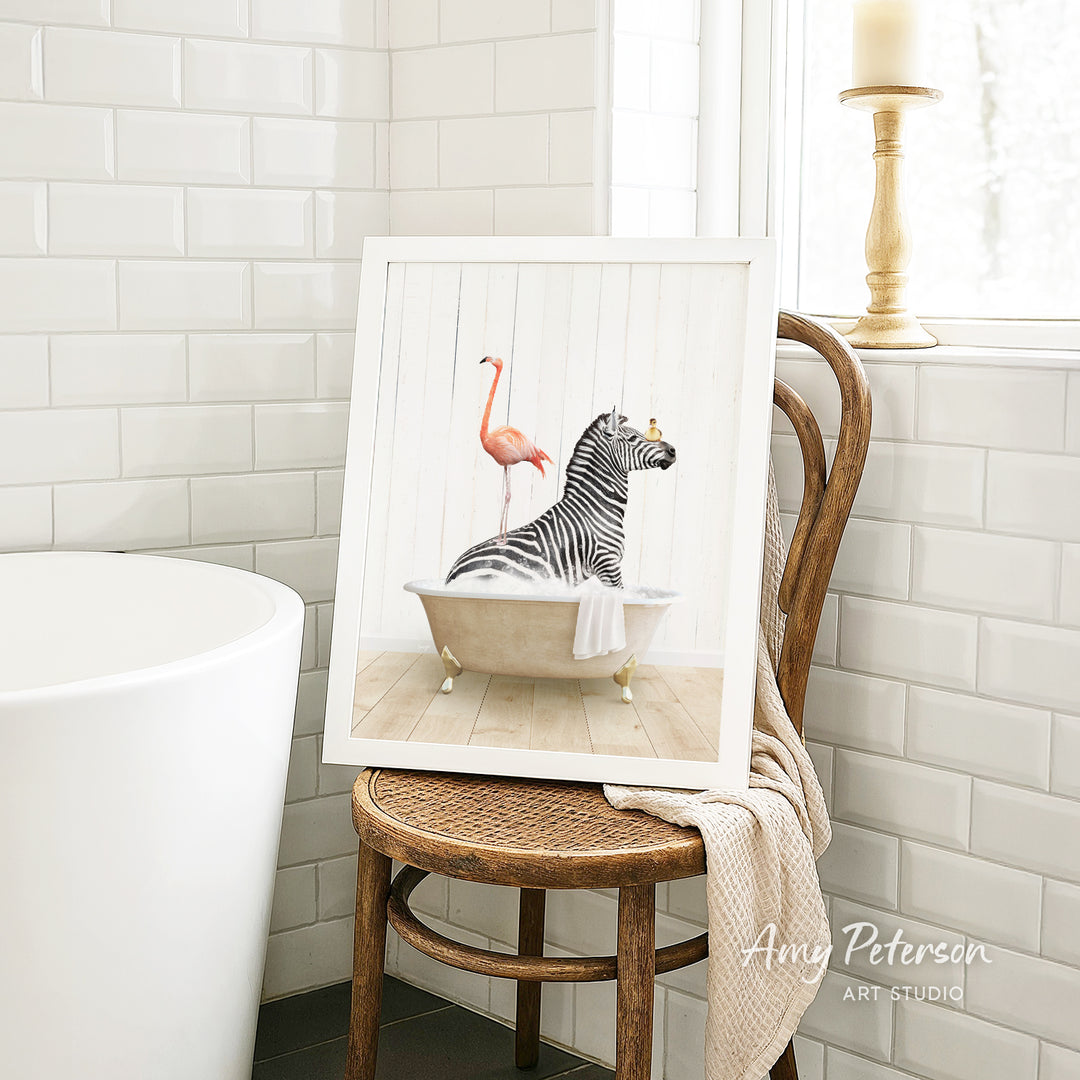 A framed image of a zebra sitting in a bathtub, with a pink flamingo standing on its back, is displayed on a wooden chair in a bathroom setting.
