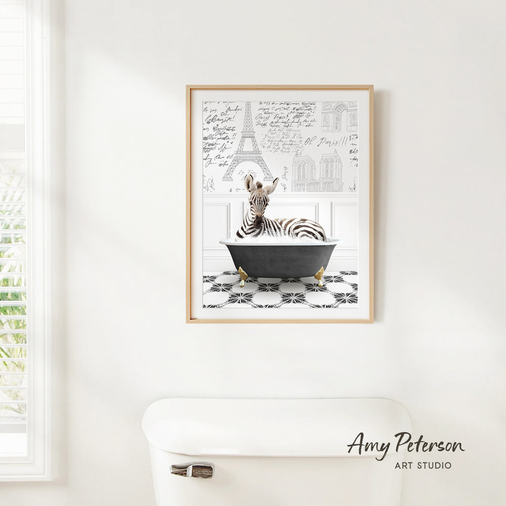 A framed print of a zebra sitting in a bathtub is hanging on a white wall, with a window and a toilet visible in the background.