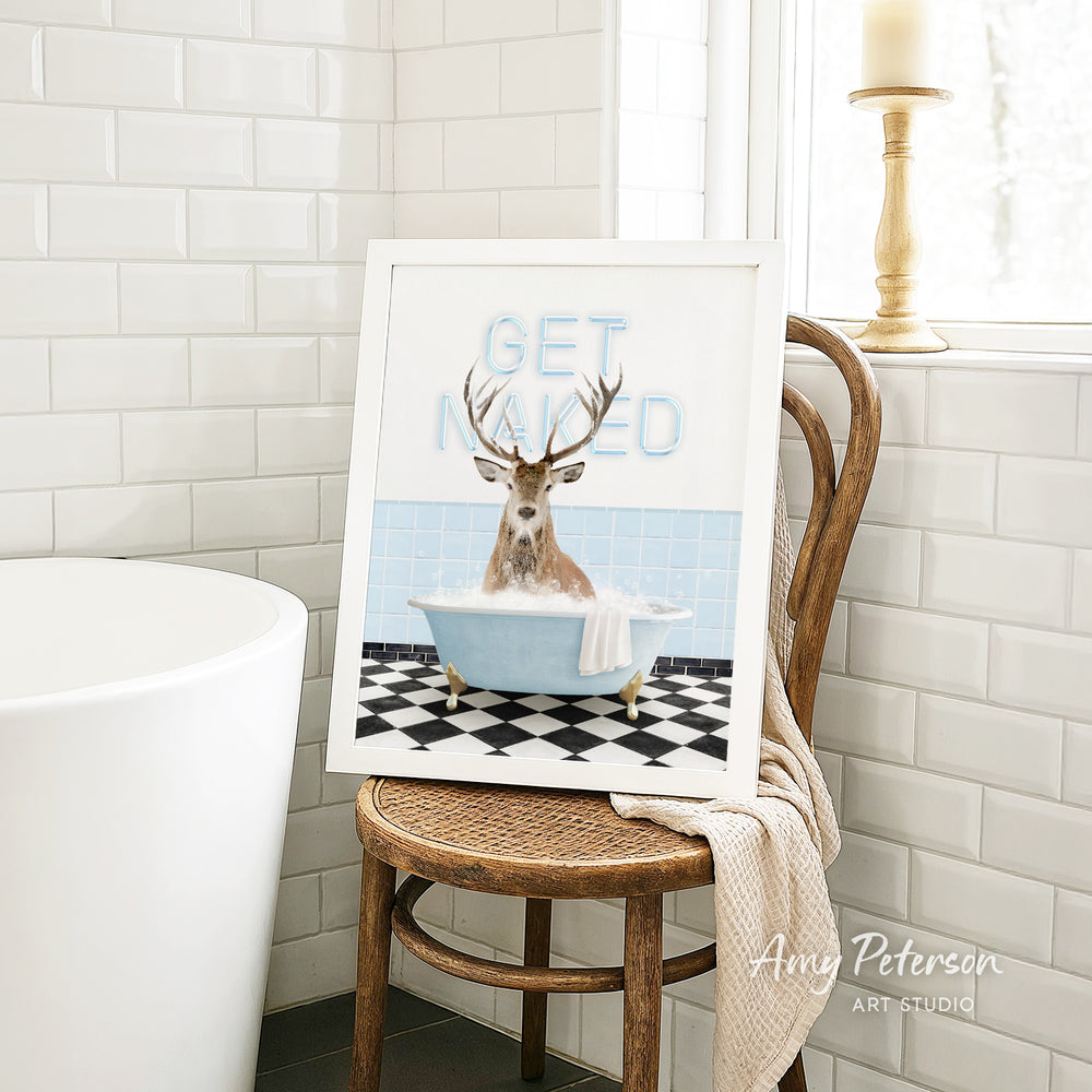 A framed image of a deer sitting in a bathtub with the text "GET NAUGED" on it, placed on a wooden chair in a bathroom setting.