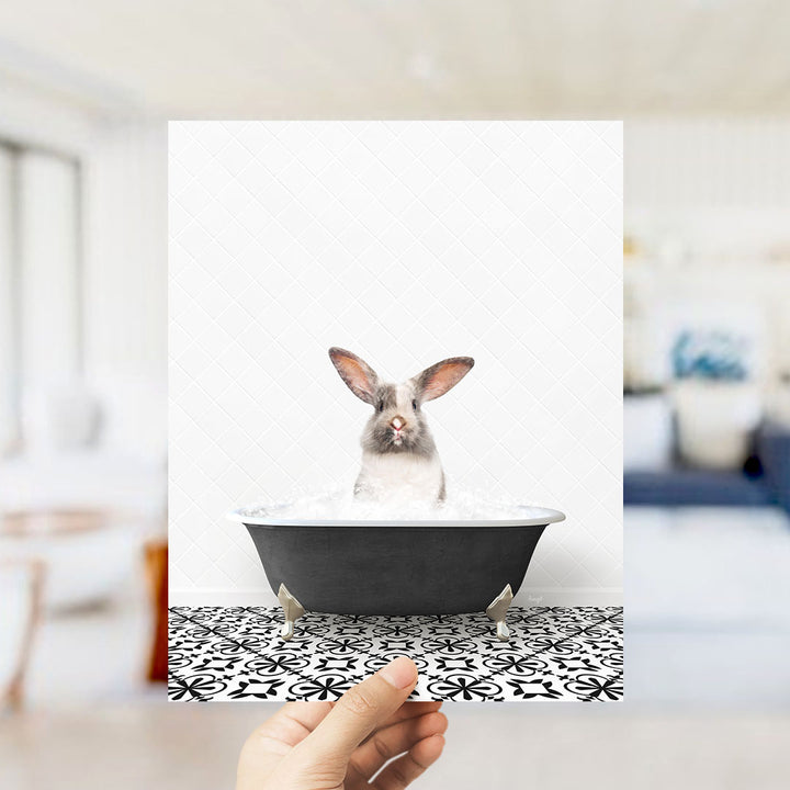 A hand holds up a framed image of a rabbit sitting in a bathtub filled with water, with a patterned tile floor visible beneath the tub.