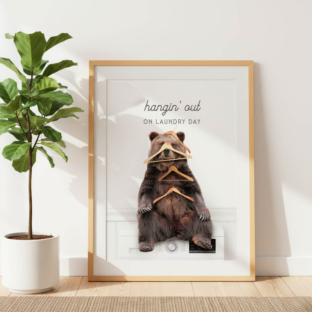 A framed image of a bear sitting on a washing machine, surrounded by hanging clothes, with the text "hangin' out on laundry day" displayed above the bear.