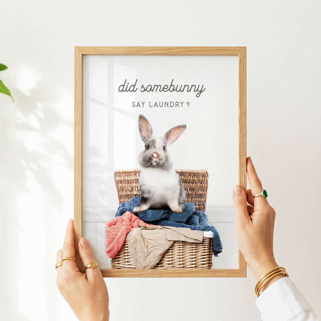 A person's hands are holding a framed picture of a rabbit sitting in a wicker basket filled with laundry.