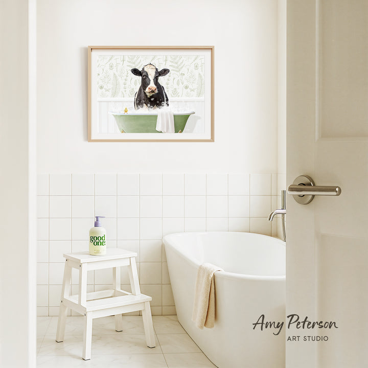 A framed image of a cow sitting in a bathtub is hanging on a white tiled wall above a white bathtub in a bathroom.