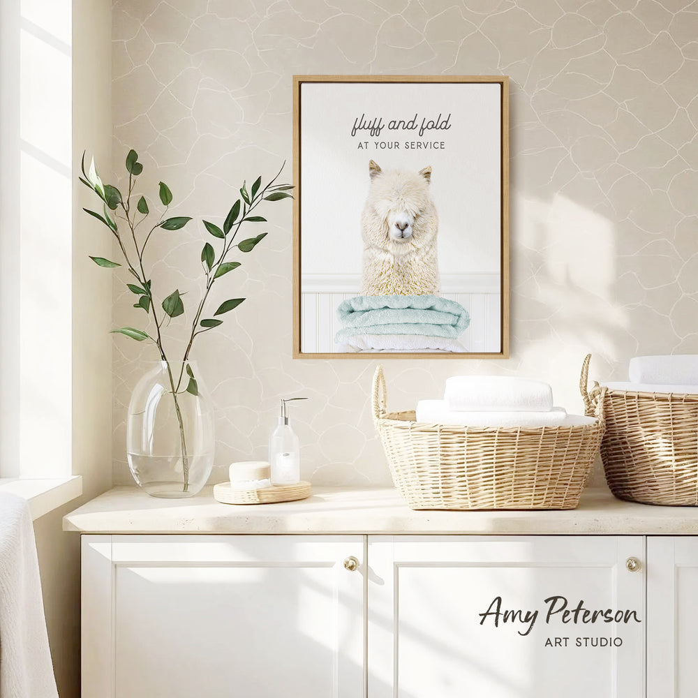 a framed picture of a fluffy alpaca sitting on a towel, hanging on a wall above a white cabinet with baskets below it.