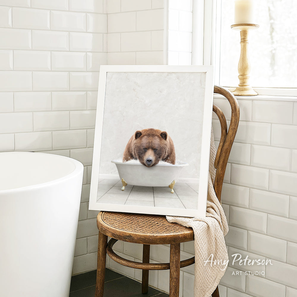 A framed image of a brown bear sitting in a white bathtub, with a towel draped over the back of a wooden chair in a bathroom setting.