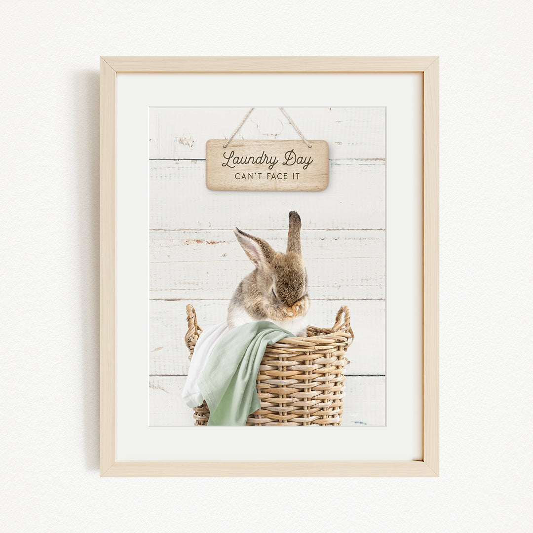 A framed image of a rabbit sitting in a wicker basket, with a wooden sign above it that says "Laundry Day Can't Face It".