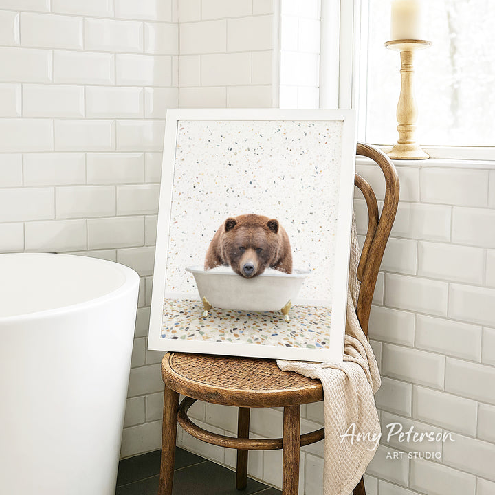 A framed image of a brown bear sitting in a bathtub is displayed on a wooden chair in a bathroom setting.