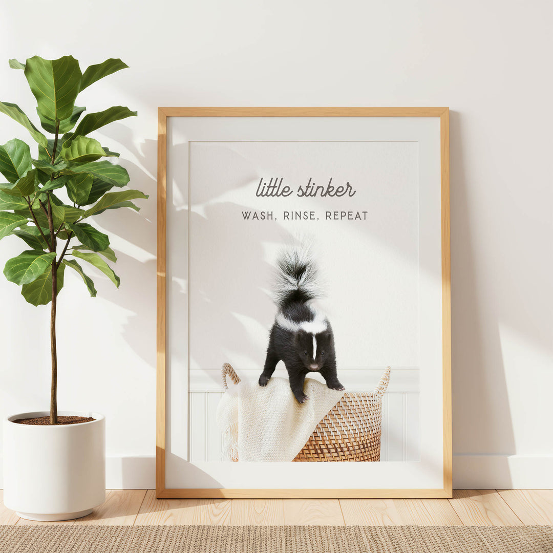 A framed poster with a skunk image and the text "little stinker" and "WASH, RINSE, REPEAT" is displayed on a white wall next to a potted plant.