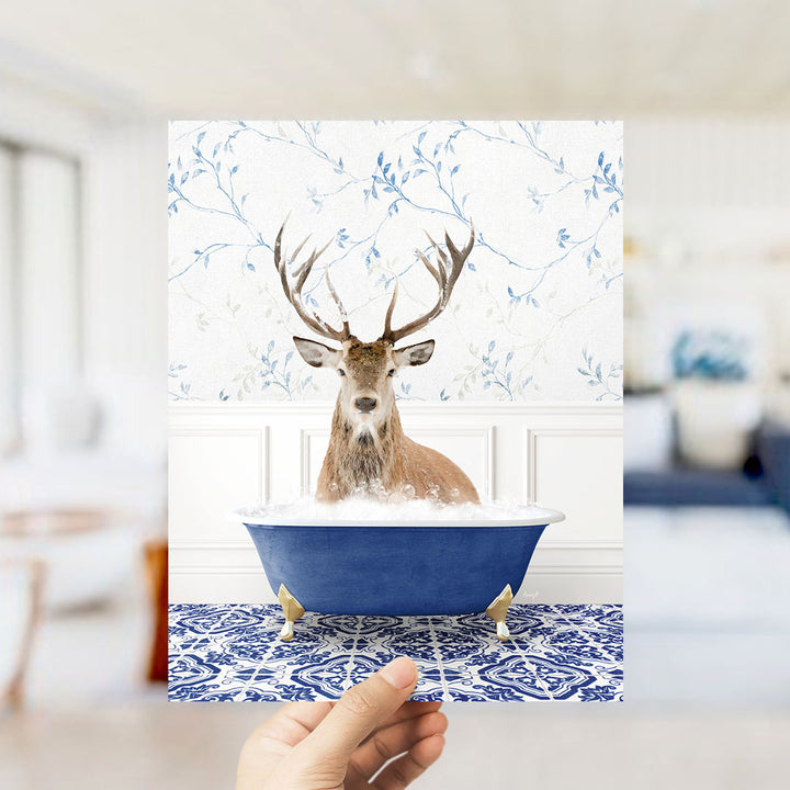 A deer with antlers is sitting in a blue bathtub filled with bubbles, set against a floral wallpaper background.