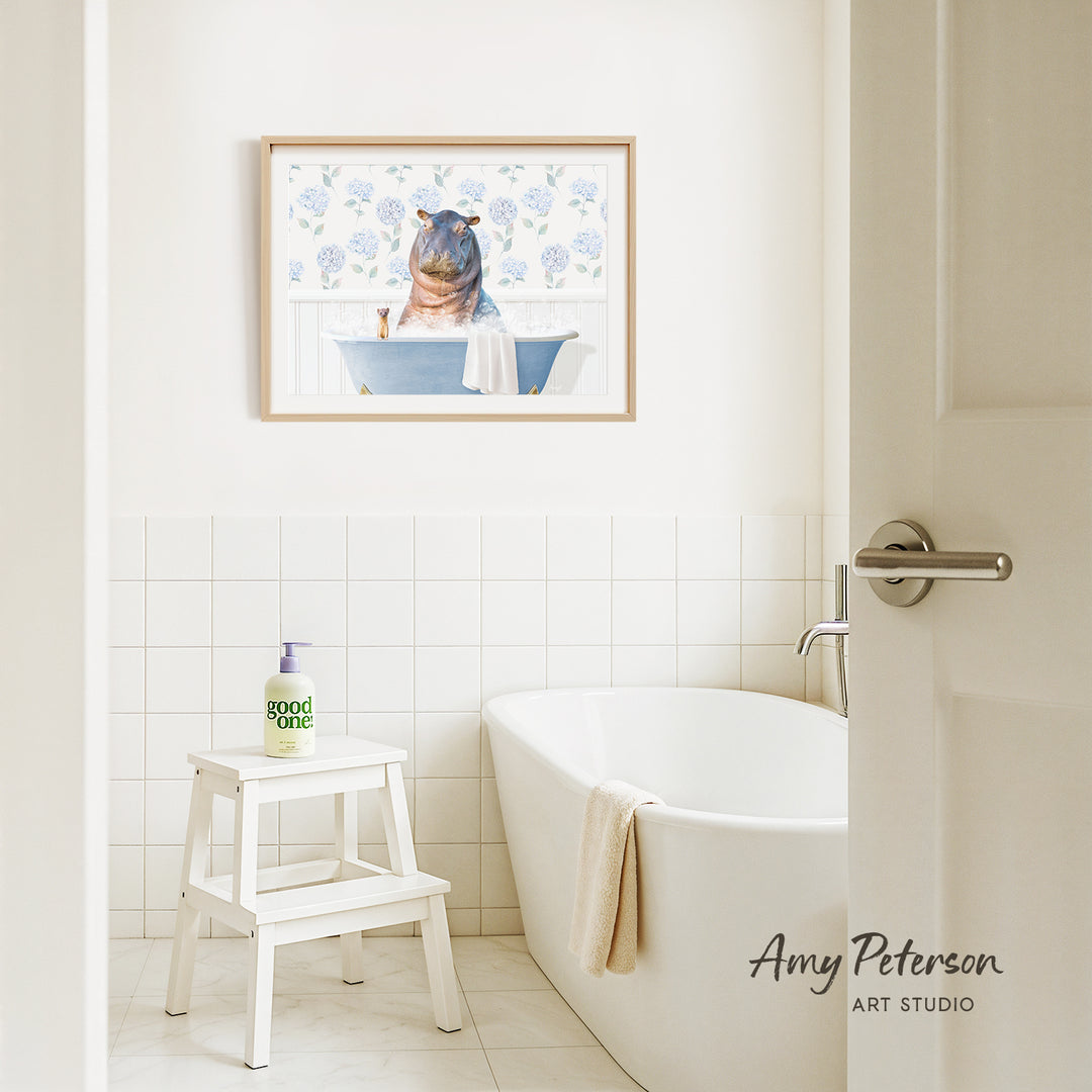 A framed painting of a hippopotamus sitting in a bathtub is hanging on a white tiled wall above a white bathtub in a bathroom.