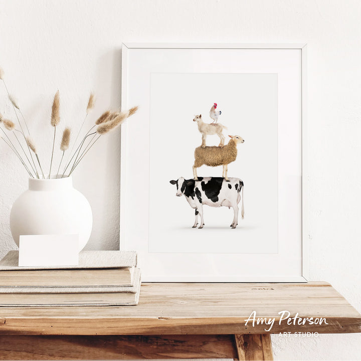 a white vase with dried reeds, a stack of books, and a framed print of a cow, sheep, and chicken on a wooden shelf.