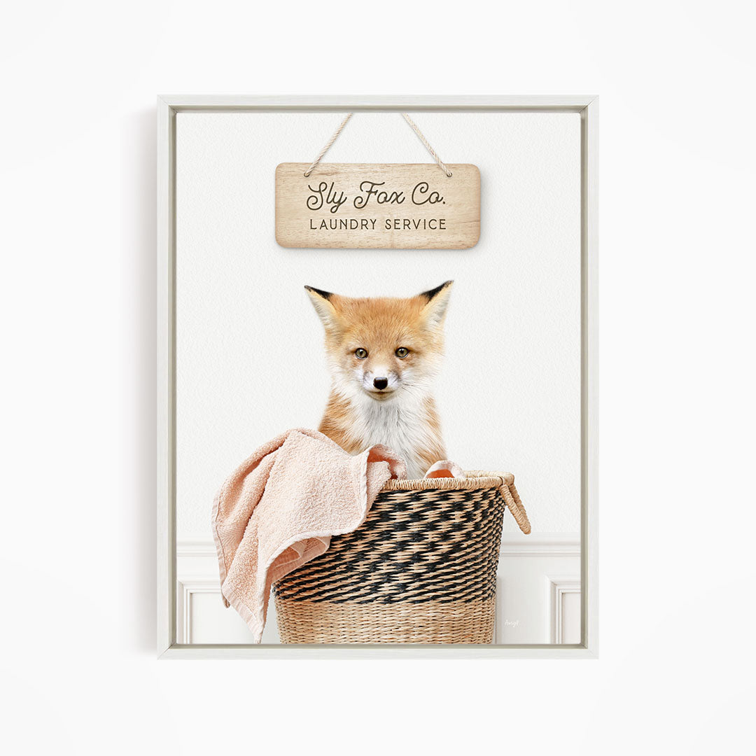 A small orange fox is sitting in a woven laundry basket, with a sign above it that reads "Fly Fox Co. Laundry Service".