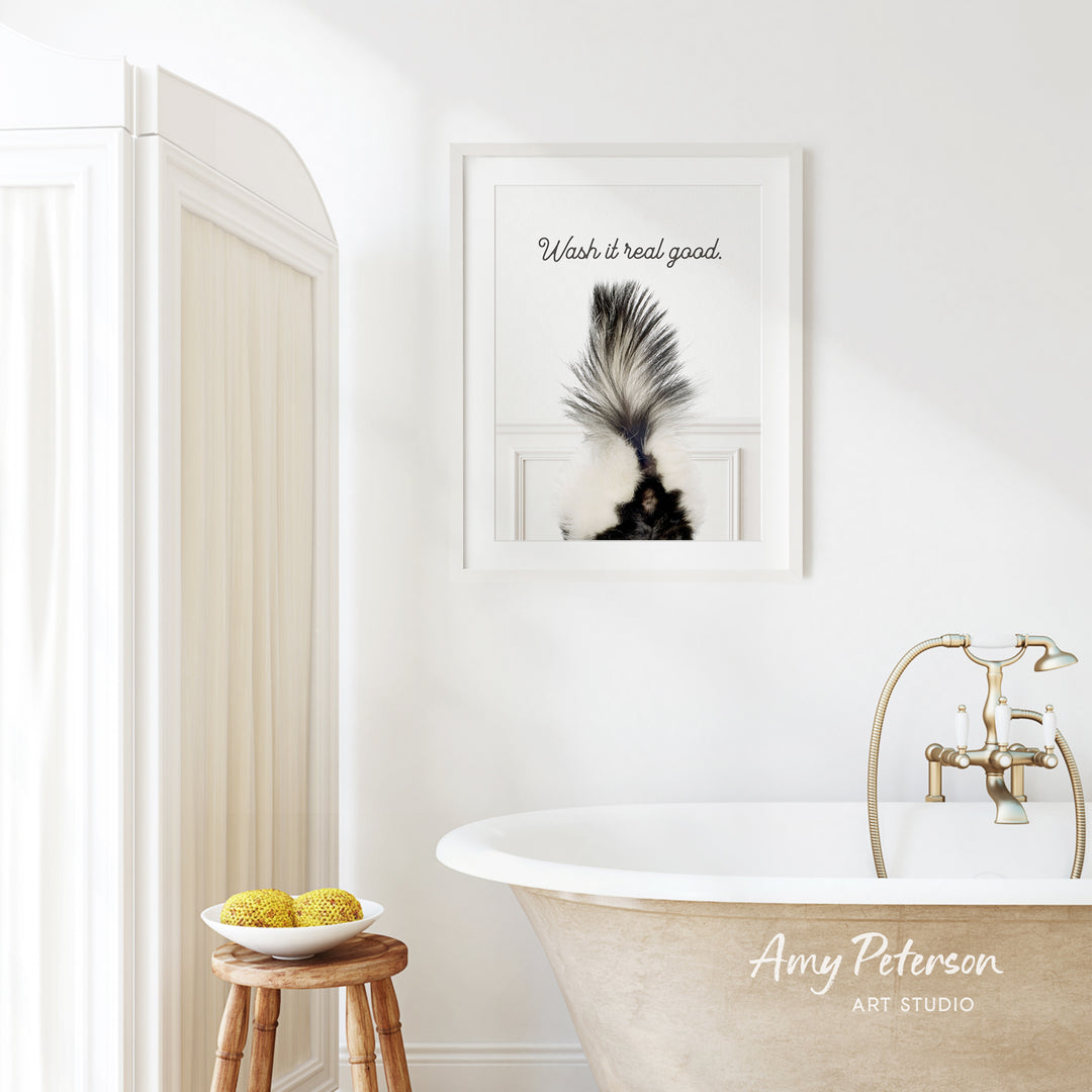 a bathroom with a framed picture of a skunk on the wall, a wooden stool with a bowl of fruit, and a clawfoot bathtub.