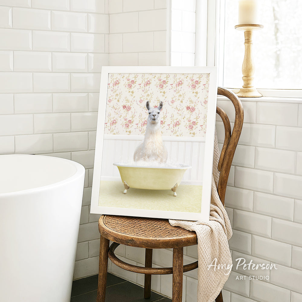 A framed image of a white llama sitting in a yellow bathtub, with a floral pattern on the wall behind it, is displayed on a wooden chair in a bathroom setting.