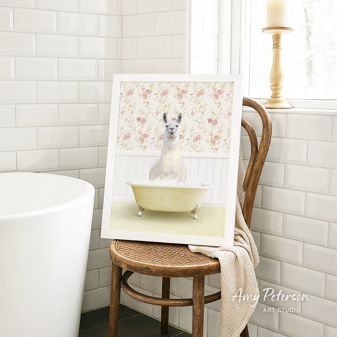 A framed image of a white llama sitting in a yellow bathtub, with a floral pattern on the wall behind it, is displayed on a wooden chair in a bathroom setting.