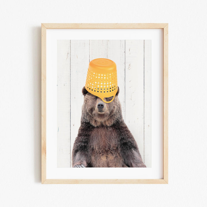 A brown bear wearing a yellow bucket hat and sitting in front of a white wooden background.