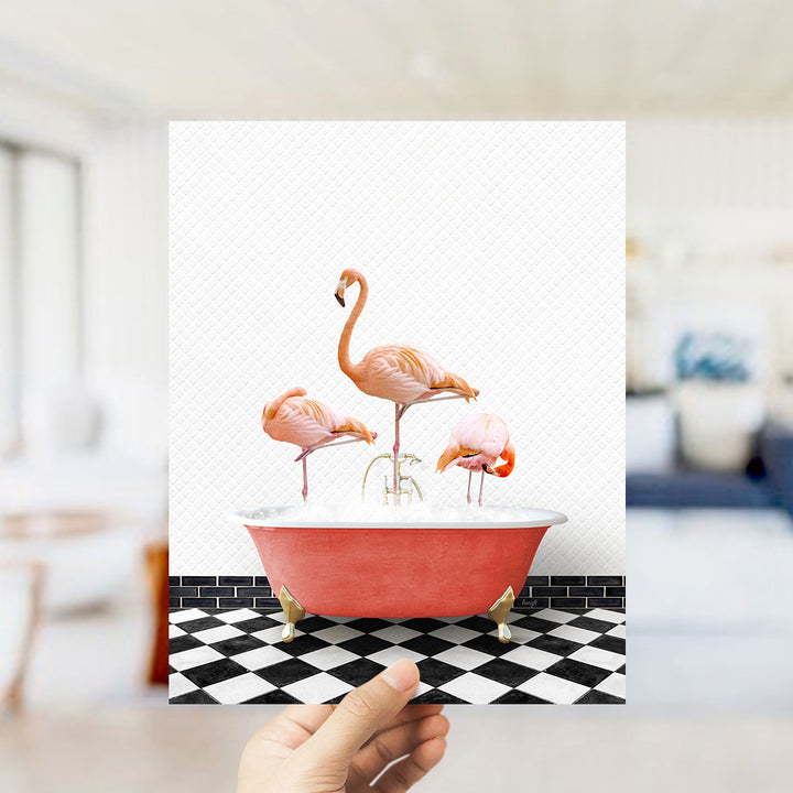 A hand holds up a framed image depicting three flamingos sitting in a red bathtub on a black and white checkered floor.