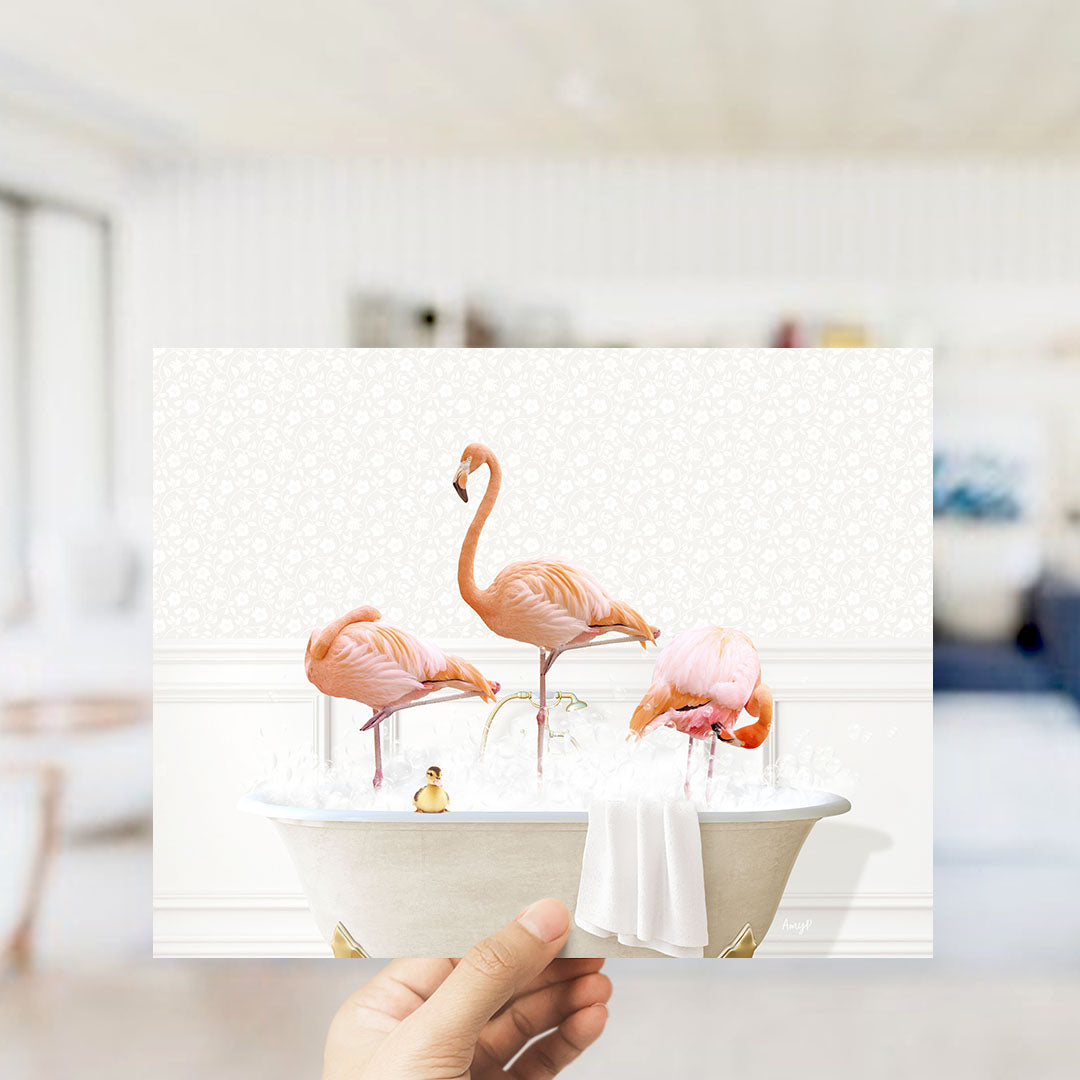 A hand holds up a framed image depicting three pink flamingos standing in a bathtub filled with water and bubbles.