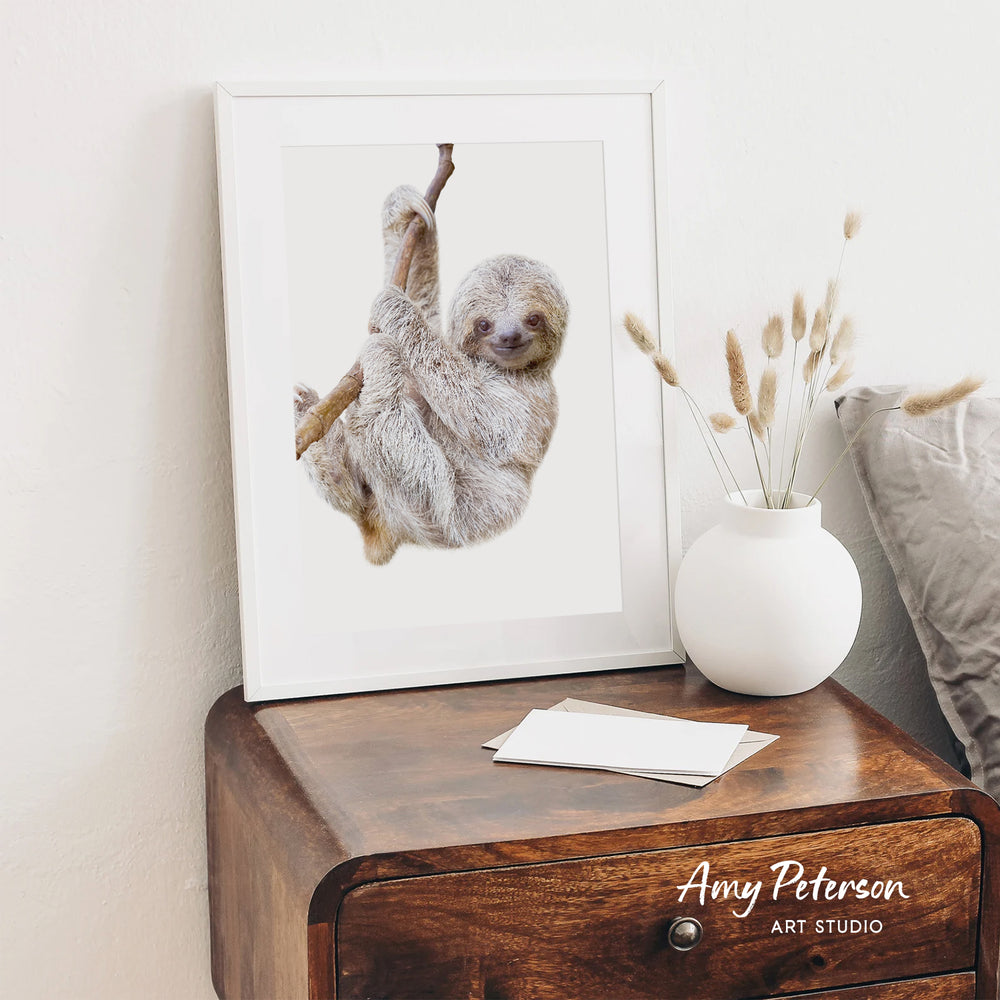 A framed image of a sloth hanging from a branch is displayed on a wooden dresser next to a white vase with dried flowers.