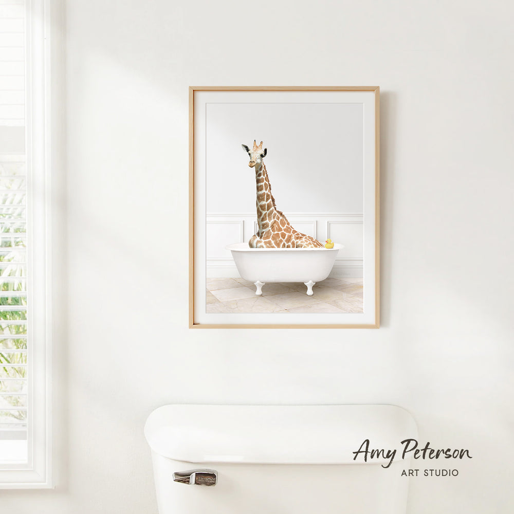 A framed image of a giraffe sitting in a white bathtub, with a window and toilet visible in the background.
