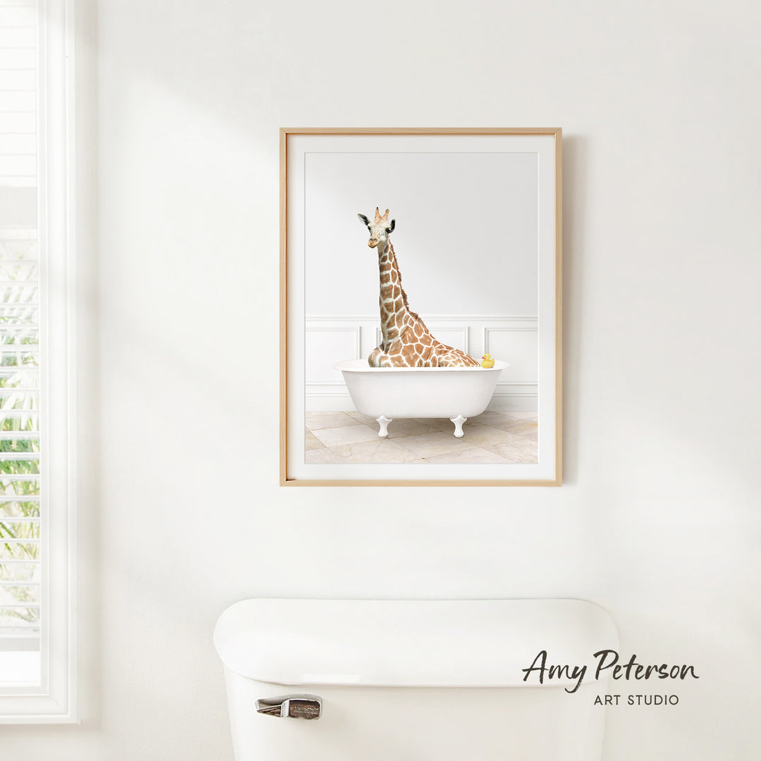 A framed image of a giraffe sitting in a white bathtub, with a window and toilet visible in the background.