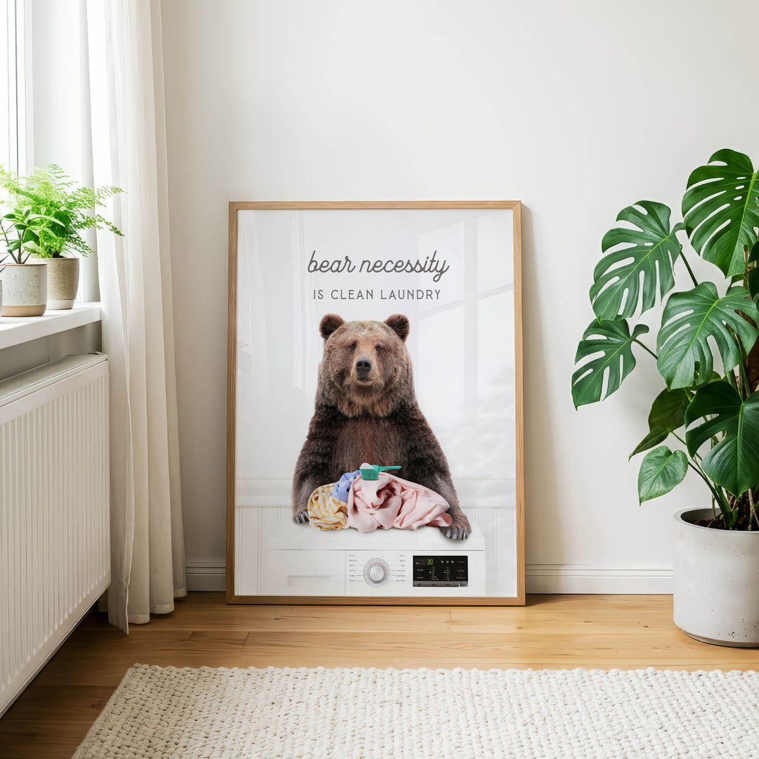 A framed poster with a brown bear sitting on a washing machine, accompanied by the text "bear necessity is clean laundry".