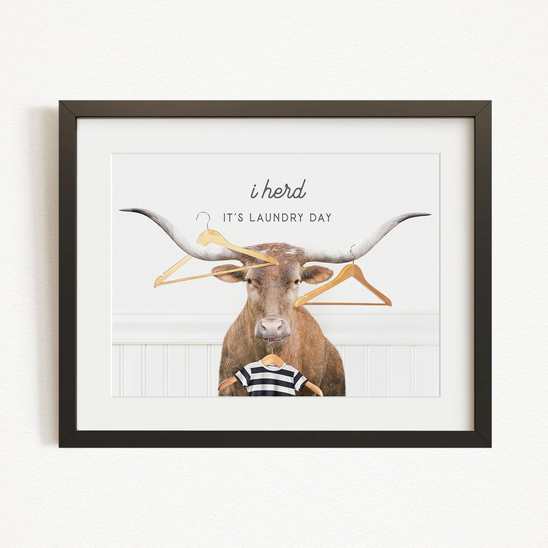 A framed image depicting a brown cow with large horns, surrounded by hanging clothes and a person wearing a striped shirt, with the text "It's laundry day" written above the cow.