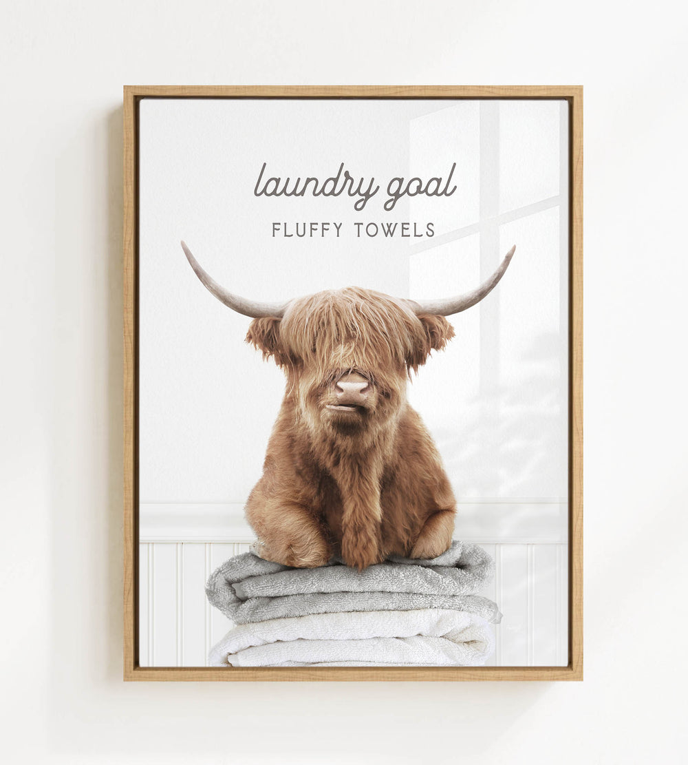 A framed image of a brown cow with large horns sitting on a stack of fluffy towels, with the text "laundry goal" and "FLUFFY TOWELS" written above it.