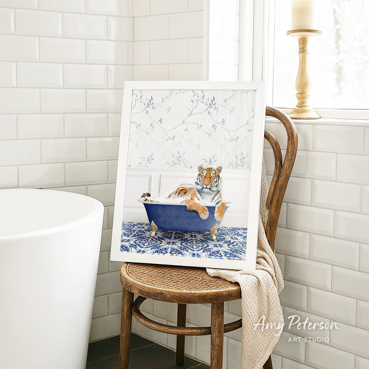 A framed painting of a tiger sitting in a blue bathtub, surrounded by a floral pattern, is displayed on a wooden chair in a bathroom setting.