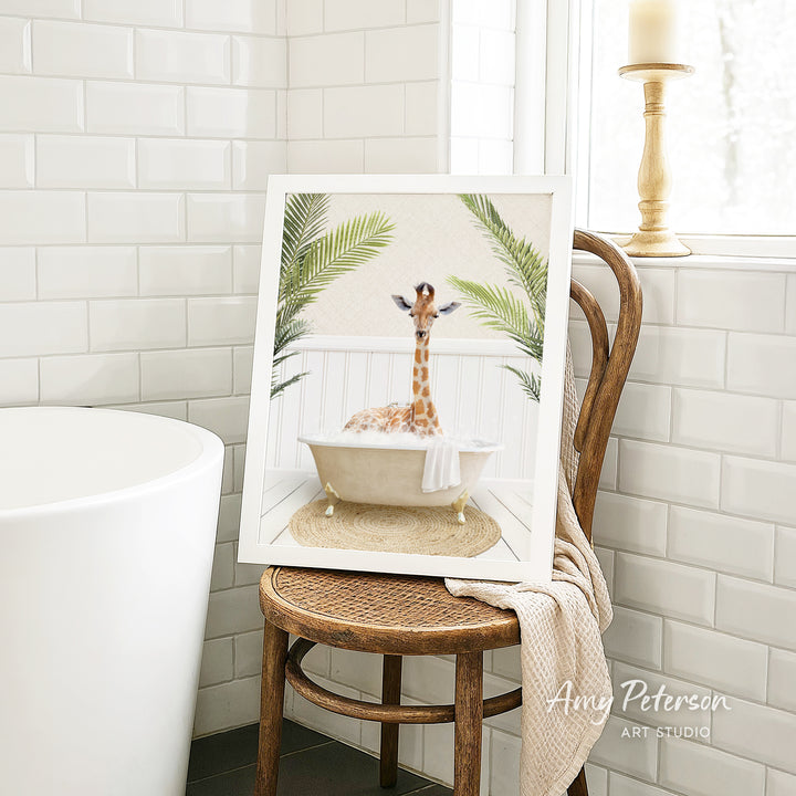 A framed image of a giraffe sitting in a bathtub, surrounded by palm leaves, is displayed on a wooden chair in a bathroom setting.