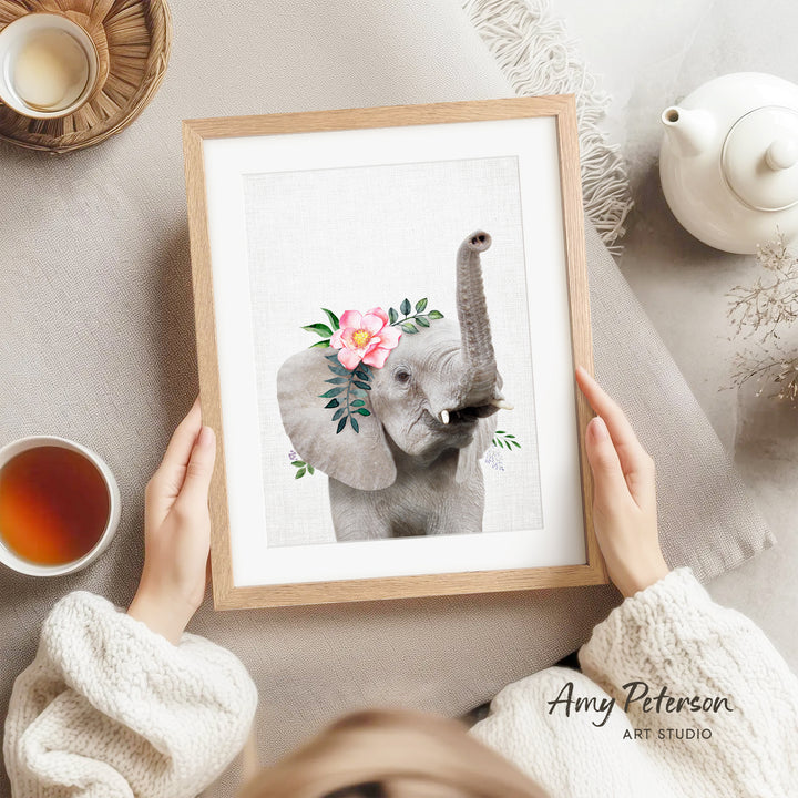 A person is holding a framed picture of an elephant with flowers on its ears, which is placed on a table. There is also a teapot, a cup, and a bowl visible in the scene.