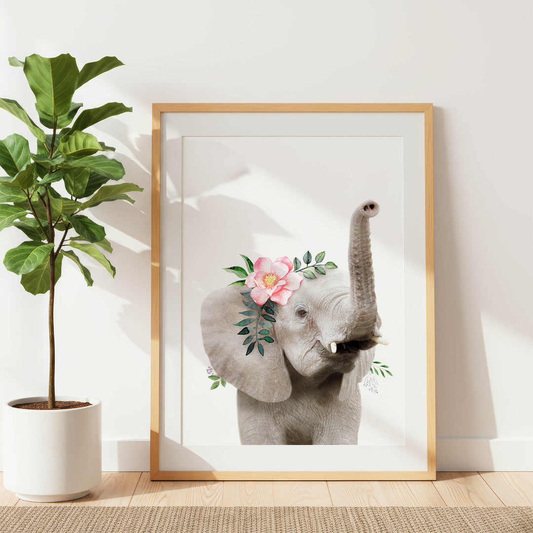 A framed print of an elephant with a pink flower on its head is displayed on a white wall, accompanied by a potted plant to the left.