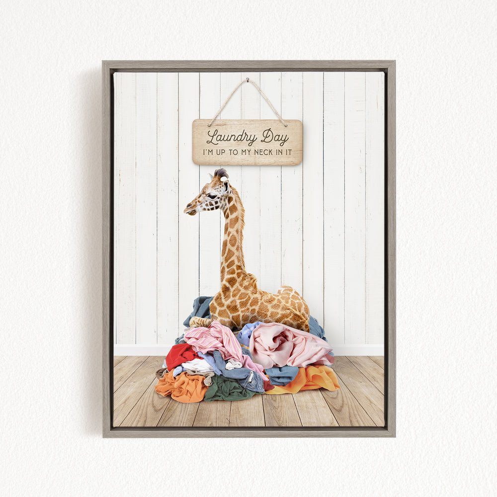 A giraffe sitting on a pile of clothes, with a wooden sign above it that says "Laundry Day I'm up to my neck in it".