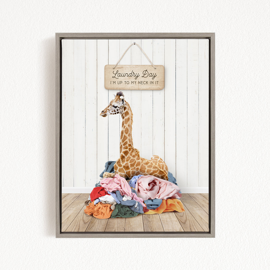 A giraffe sitting on a pile of clothes, with a wooden sign above it that says "Laundry Day I'm up to my neck in it".