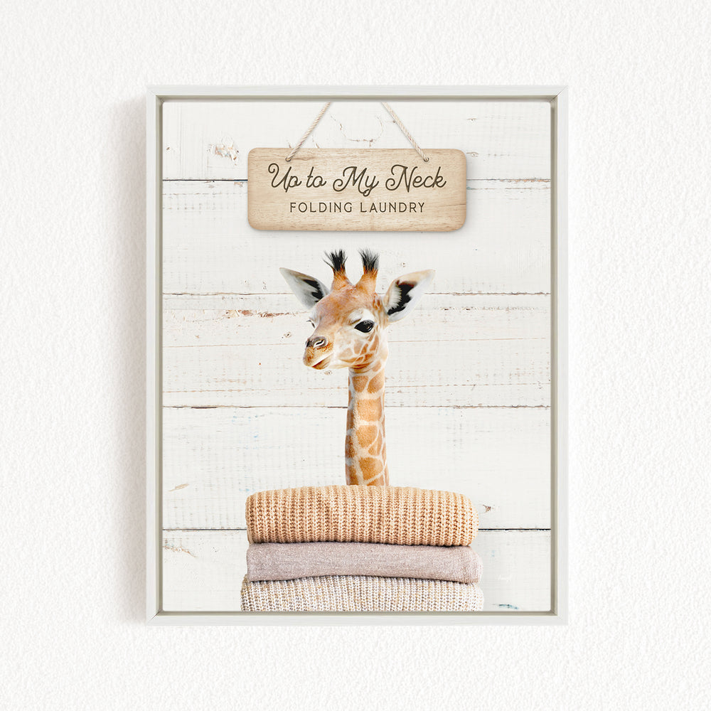A framed image of a giraffe standing on a pile of folded clothes, with a wooden sign above it that says "Up to My Neck Folding Laundry".