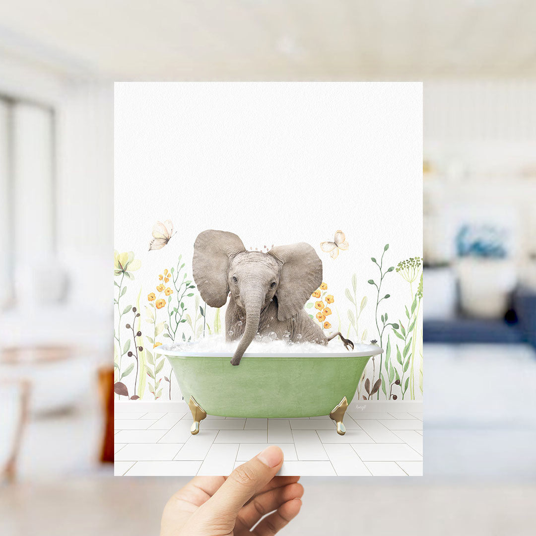 A hand holds up a framed image depicting a baby elephant sitting in a green bathtub filled with bubbles, surrounded by flowers and butterflies.