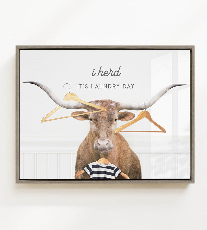 A framed image depicting a bull with a child wearing a striped shirt, surrounded by clothes hangers, with the text "i herd, it's laundry day" above the bull.