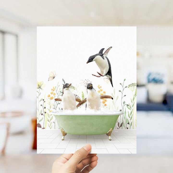A hand holds up a framed image depicting two penguins sitting in a green bathtub, surrounded by flowers and plants.