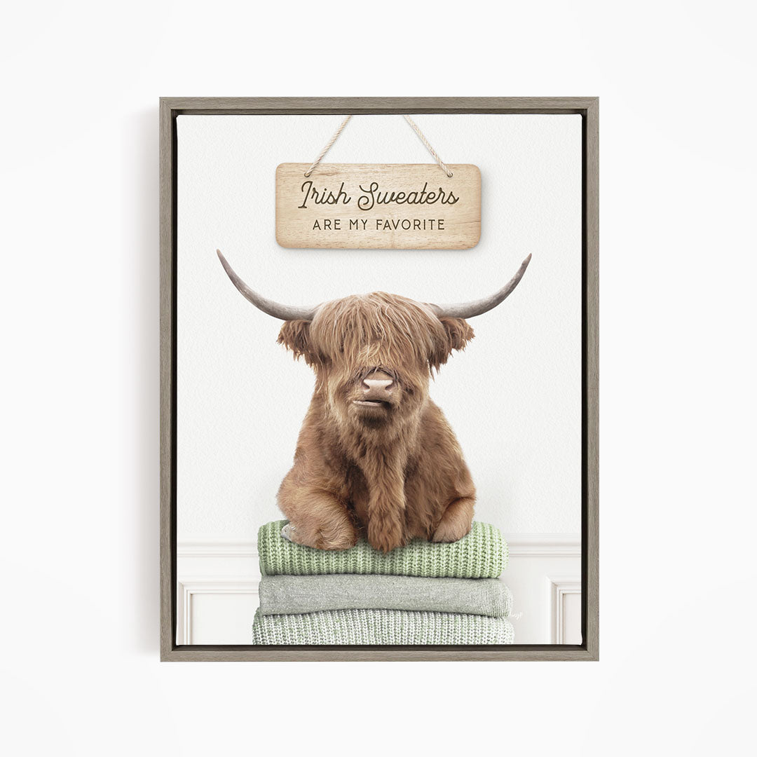 A brown Highland cow with large horns is sitting on a stack of green and gray blankets, with a wooden sign above that says "Irish Sweaters are my favorite".