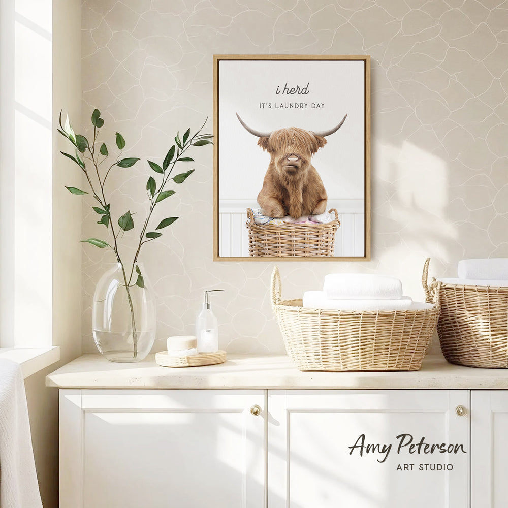 a framed poster of a Highland cow sitting in a laundry basket, with the caption "It's laundry day" above it. The poster is displayed on a wall above a white cabinet, which has a vase of leaves and a bottle of soap on it.