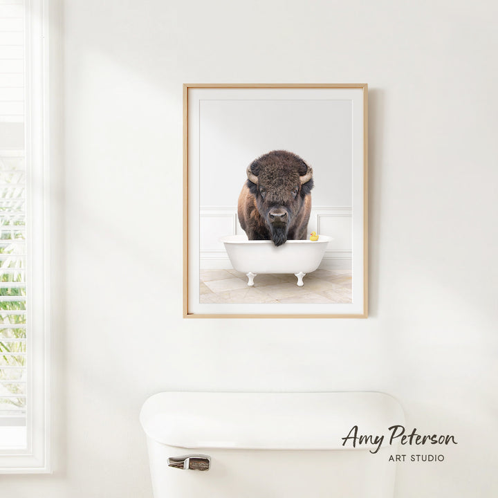 A framed image of a buffalo standing in a white bathtub, with a window and toilet visible in the background.
