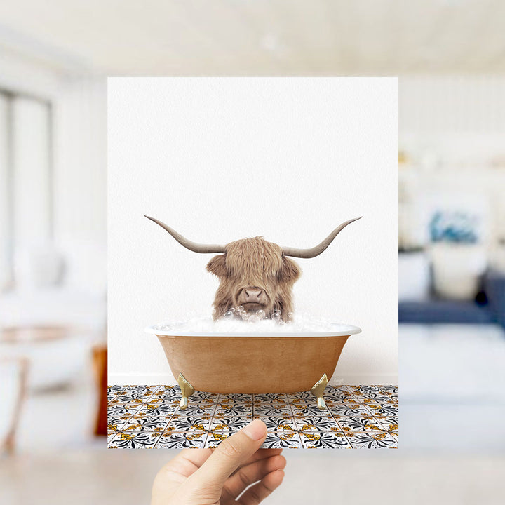 A hand holds up a framed image of a Highland cow with large horns sitting in a bathtub filled with water.
