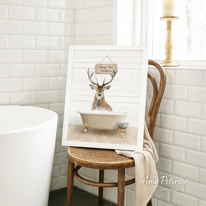 A framed image of a deer sitting in a bathtub, with a sign above it that says "Wish Your Antlers". The image is displayed on a wooden chair in a bathroom setting.