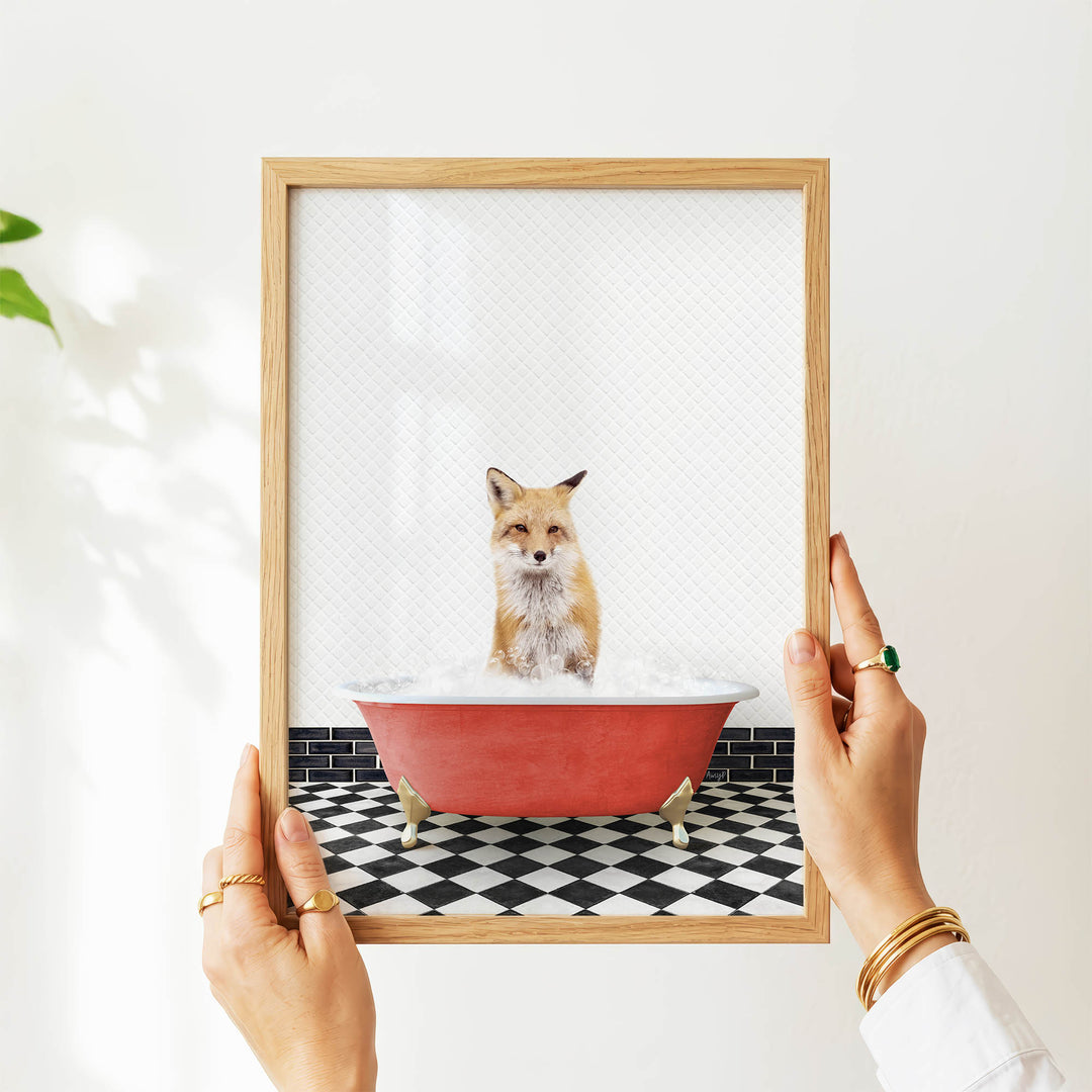 A person's hands are holding a framed picture of a fox sitting in a red bathtub filled with water, with a black and white checkered floor visible beneath the tub.