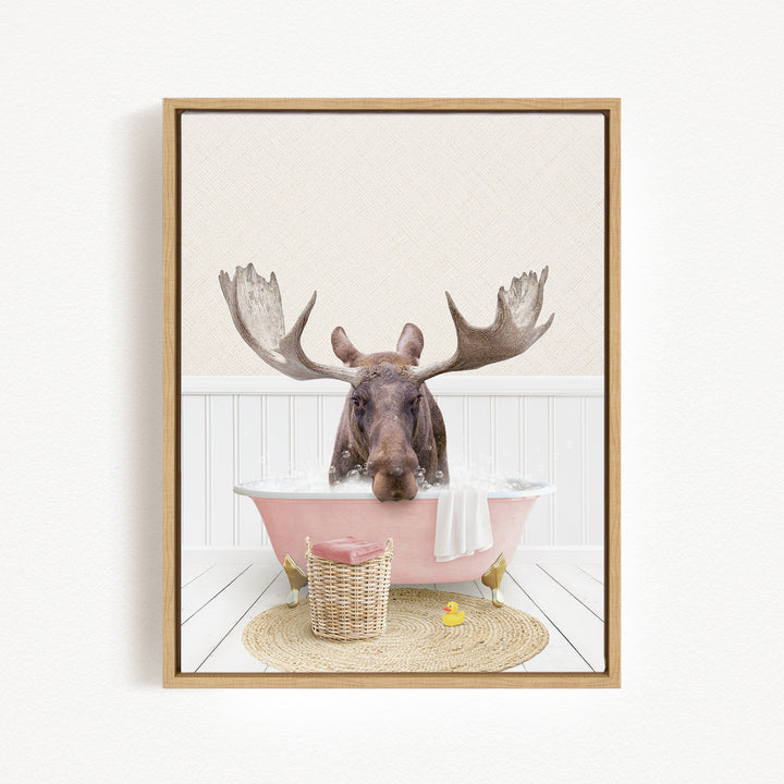 A moose with large antlers is sitting in a pink bathtub filled with water, surrounded by a basket of toys and a yellow rubber duck.