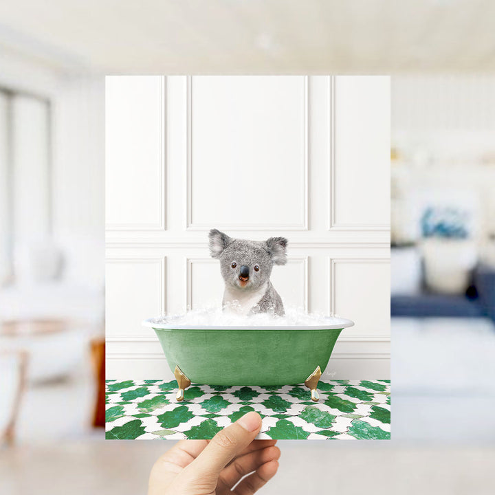 A hand holds up a framed image of a koala bear sitting in a green bathtub filled with bubbles, surrounded by a green and white patterned floor.