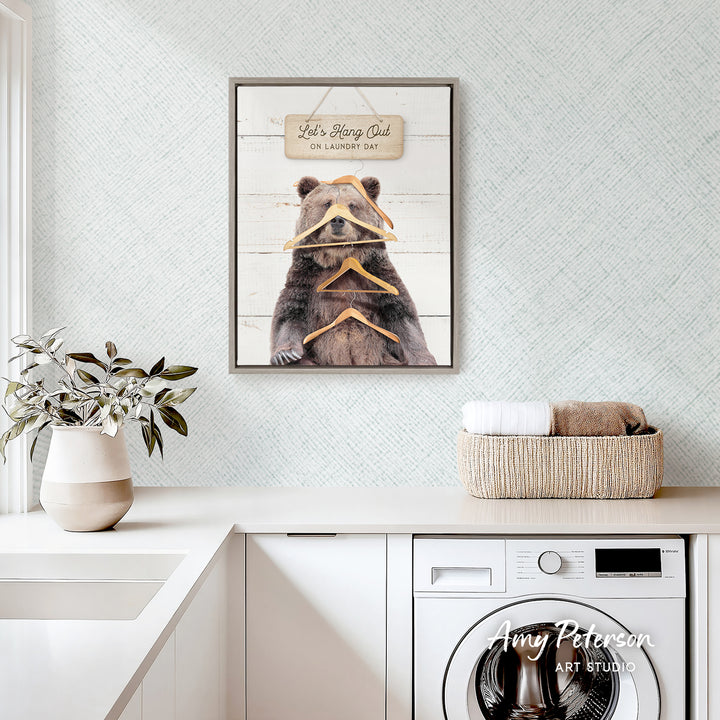 a framed picture of a bear wearing a sign that says "Let's Hang Out on Laundry Day" above a white sink and a basket of towels.