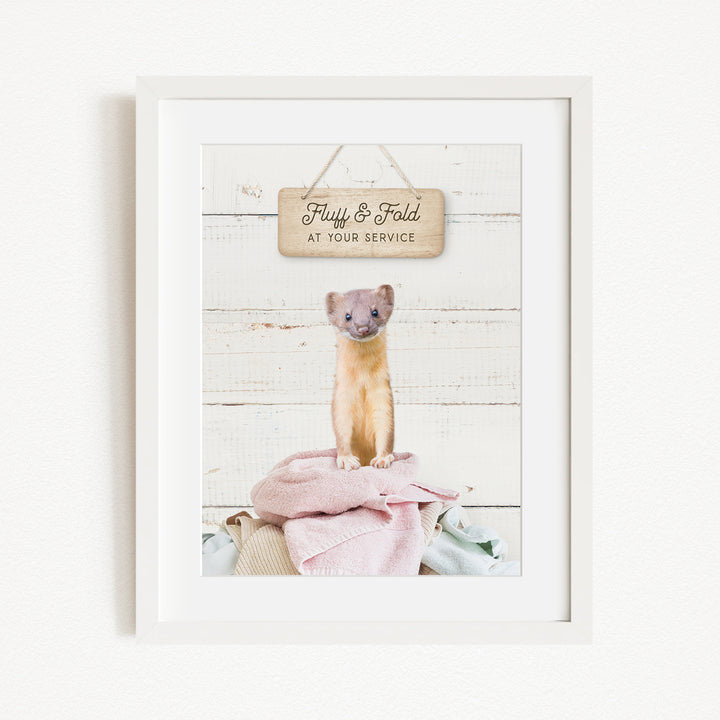A framed image of a small animal, likely a cat, sitting on a pile of clothes and towels, with a wooden sign above it that says "Fluffy & Fold at your service".