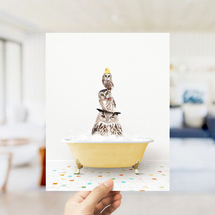 A hand holds up a framed image depicting three owls sitting in a yellow bathtub filled with bubbles.